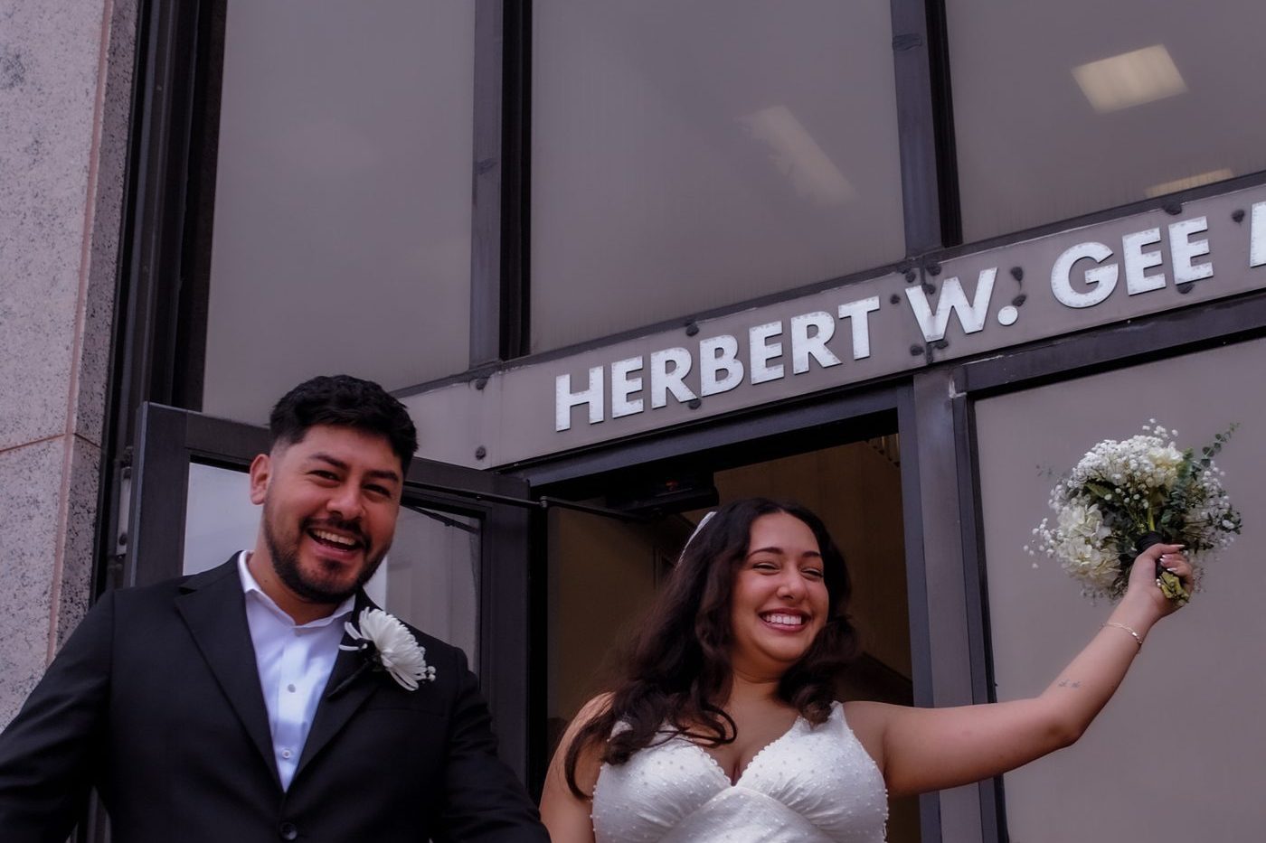 Joyful newlyweds exit the courthouse hand in hand, bouquet raised